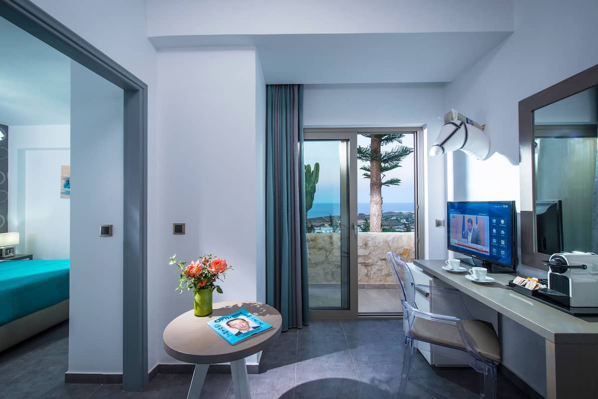 A modern hotel room with a balcony, a desk with a computer, a coffee maker, and a vase with flowers.