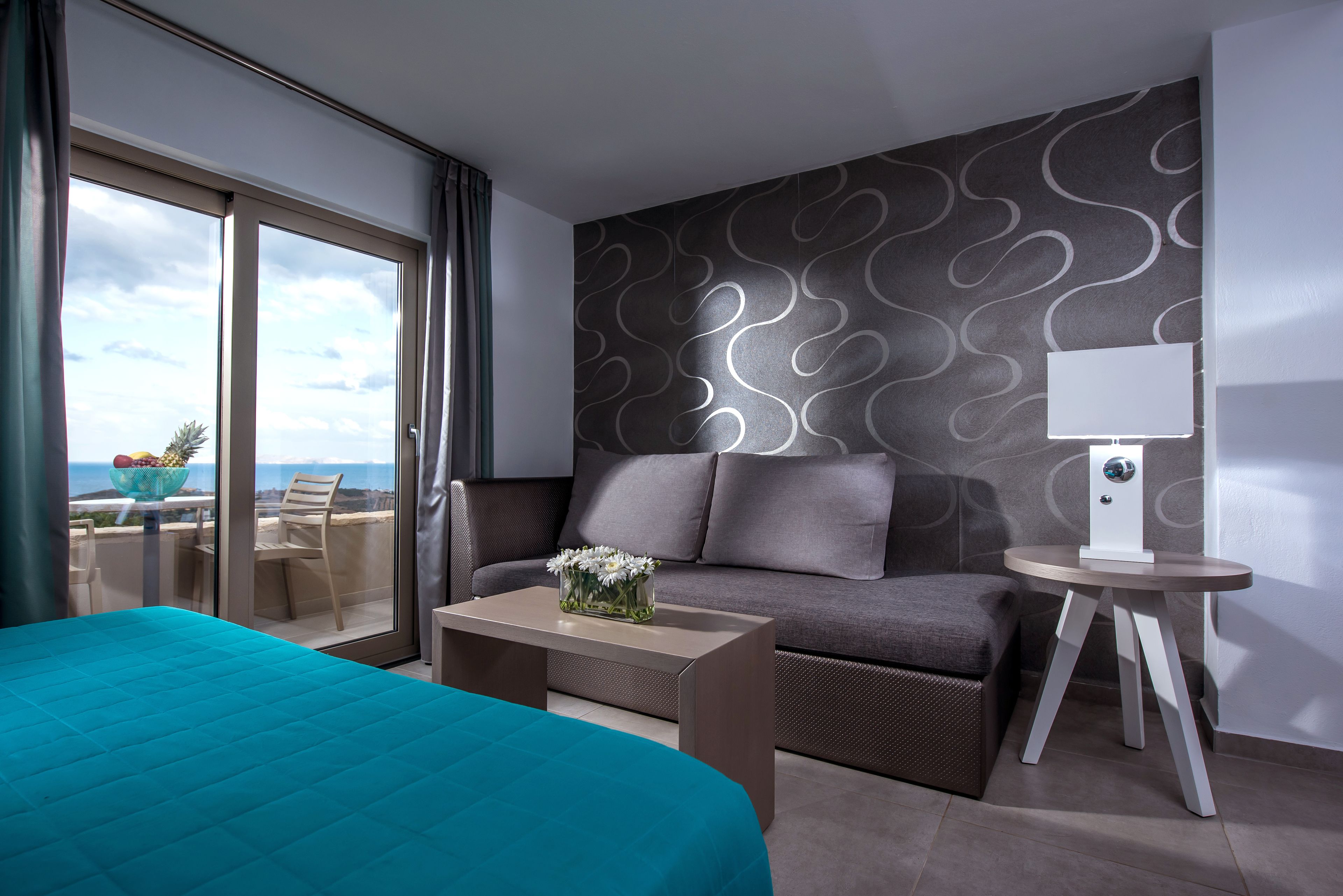 Ein Hotelzimmer mit Bett, Sofa, Couchtisch, einem kleinen Tisch mit Lampe sowie einem Balkon mit Blick auf die Stadt und das Meer.