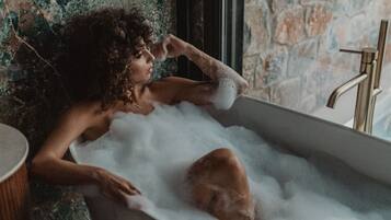 Una persona relajándose en una bañera llena de burbujas, con una pared de piedra y una ventana al fondo.