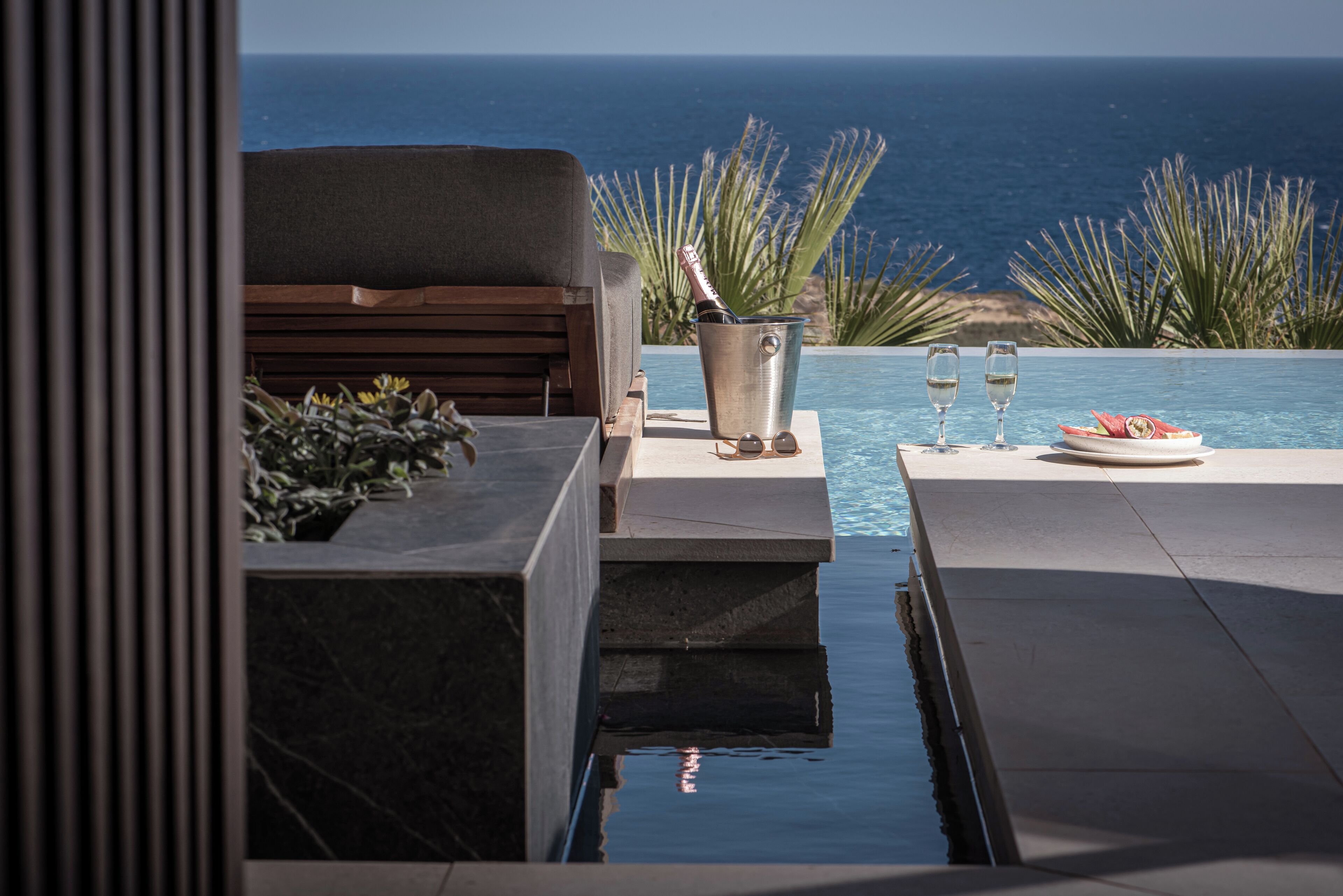Un espace aménagé au bord de la piscine, avec vue sur l’océan, un banc en bois, un seau en métal contenant une bouteille et deux verres posés sur une table.