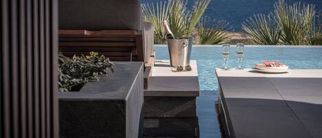 Un espace aménagé au bord de la piscine, avec vue sur l’océan, un banc en bois, un seau en métal contenant une bouteille et deux verres posés sur une table.