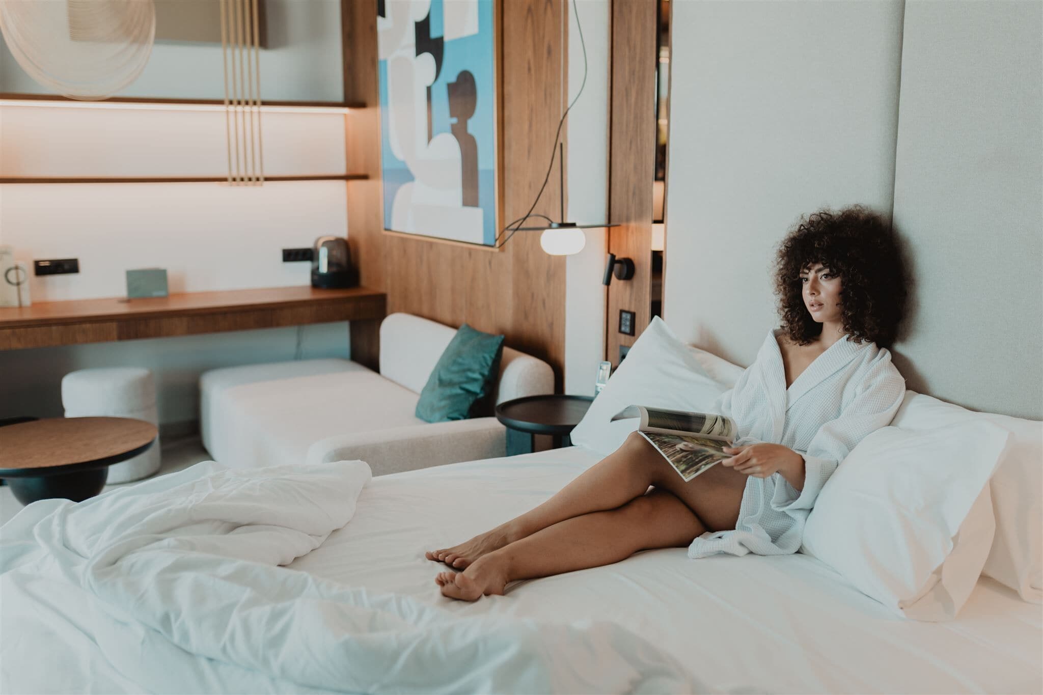A modern hotel room with a bed, a bedside table, a reading lamp, and a wall-mounted artwork.