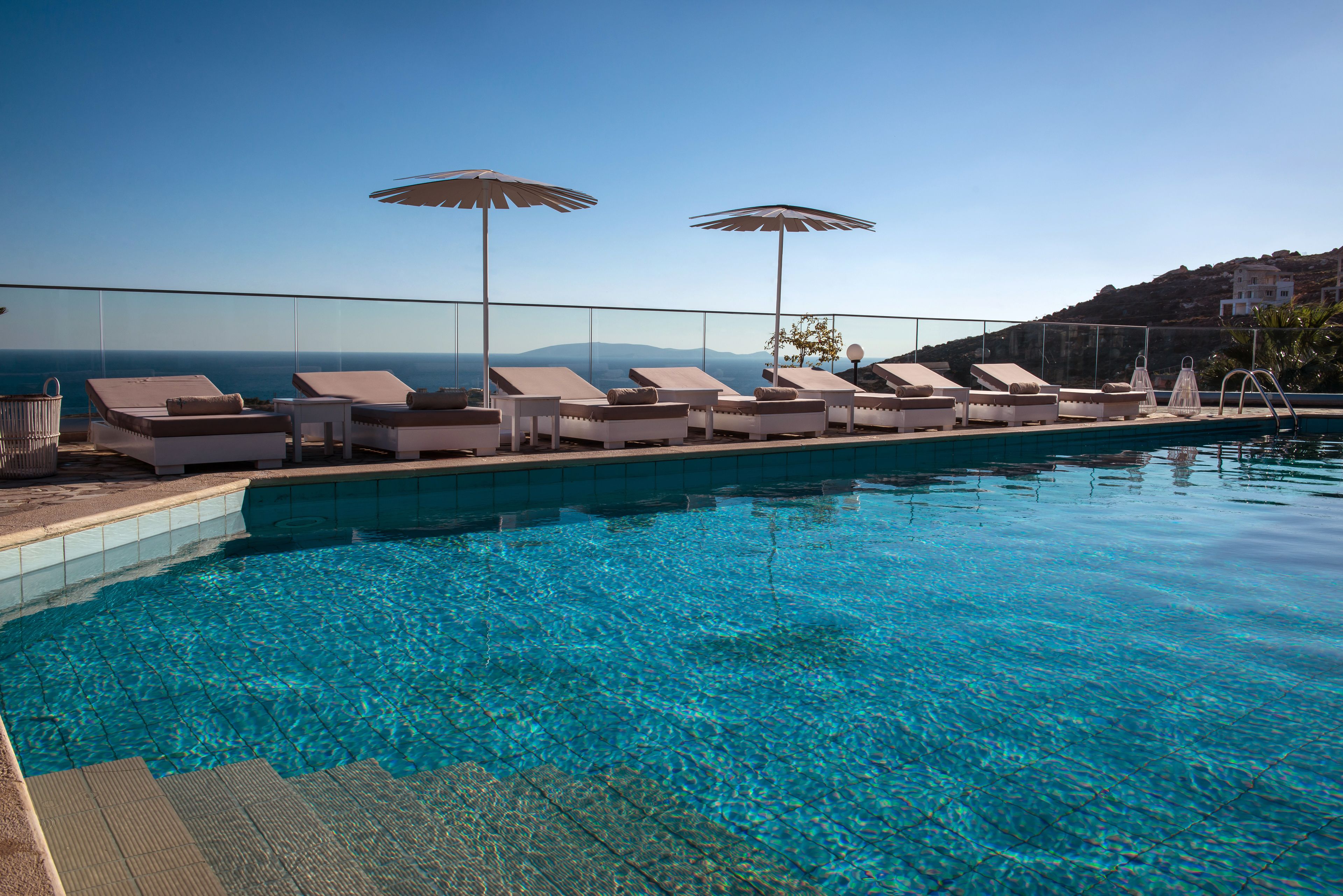 Zona junto a la piscina con sillones de descanso y sombrillas, agua azul cristalina y vistas al mar y colinas.