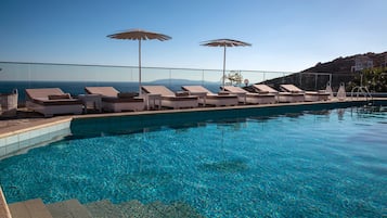 Zona junto a la piscina con sillones de descanso y sombrillas, agua azul cristalina y vistas al mar y colinas.