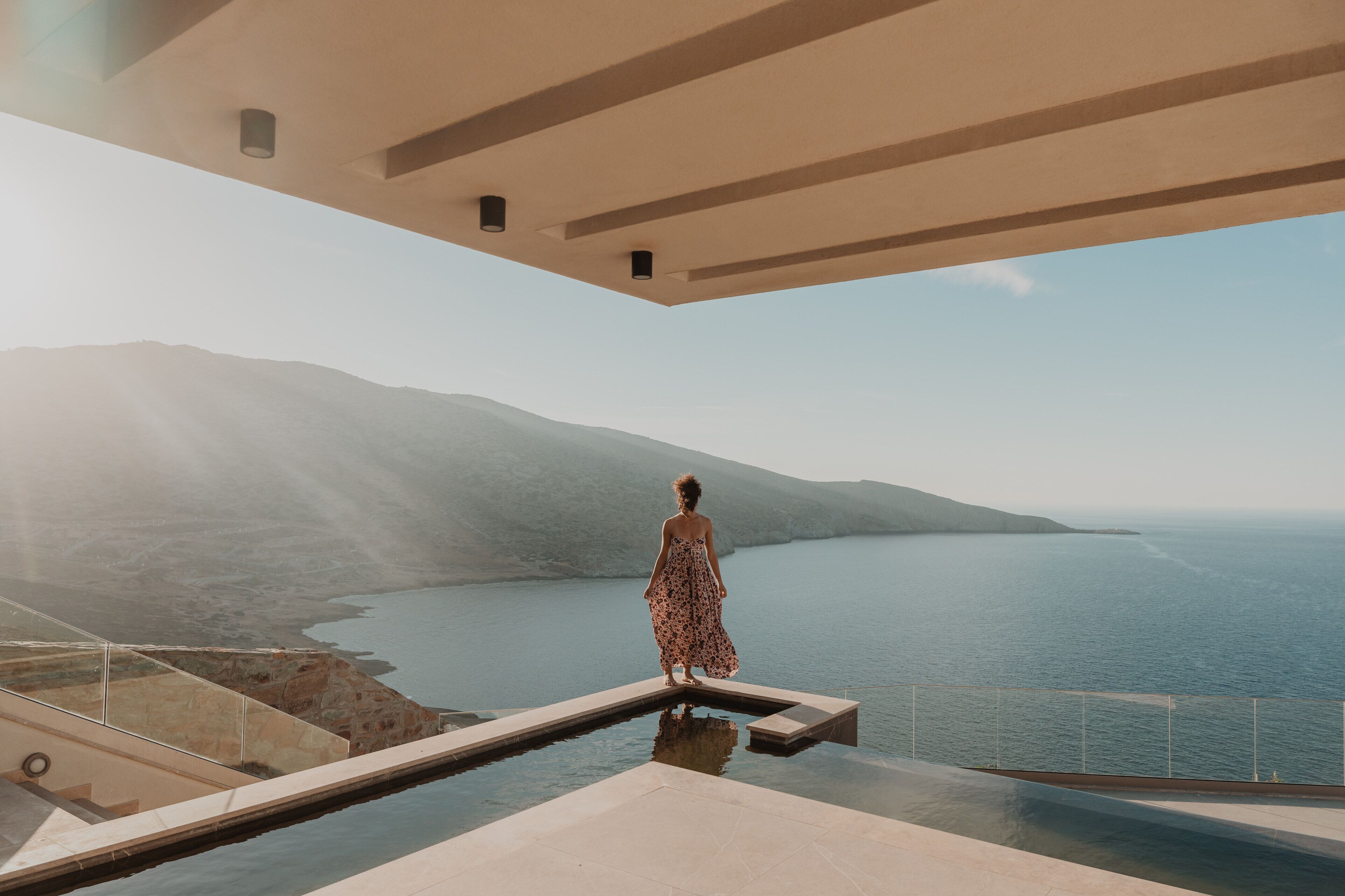 Une femme se tient sur un balcon, d’où elle a une vue sur une étendue d’eau, avec des montagnes en arrière-plan.