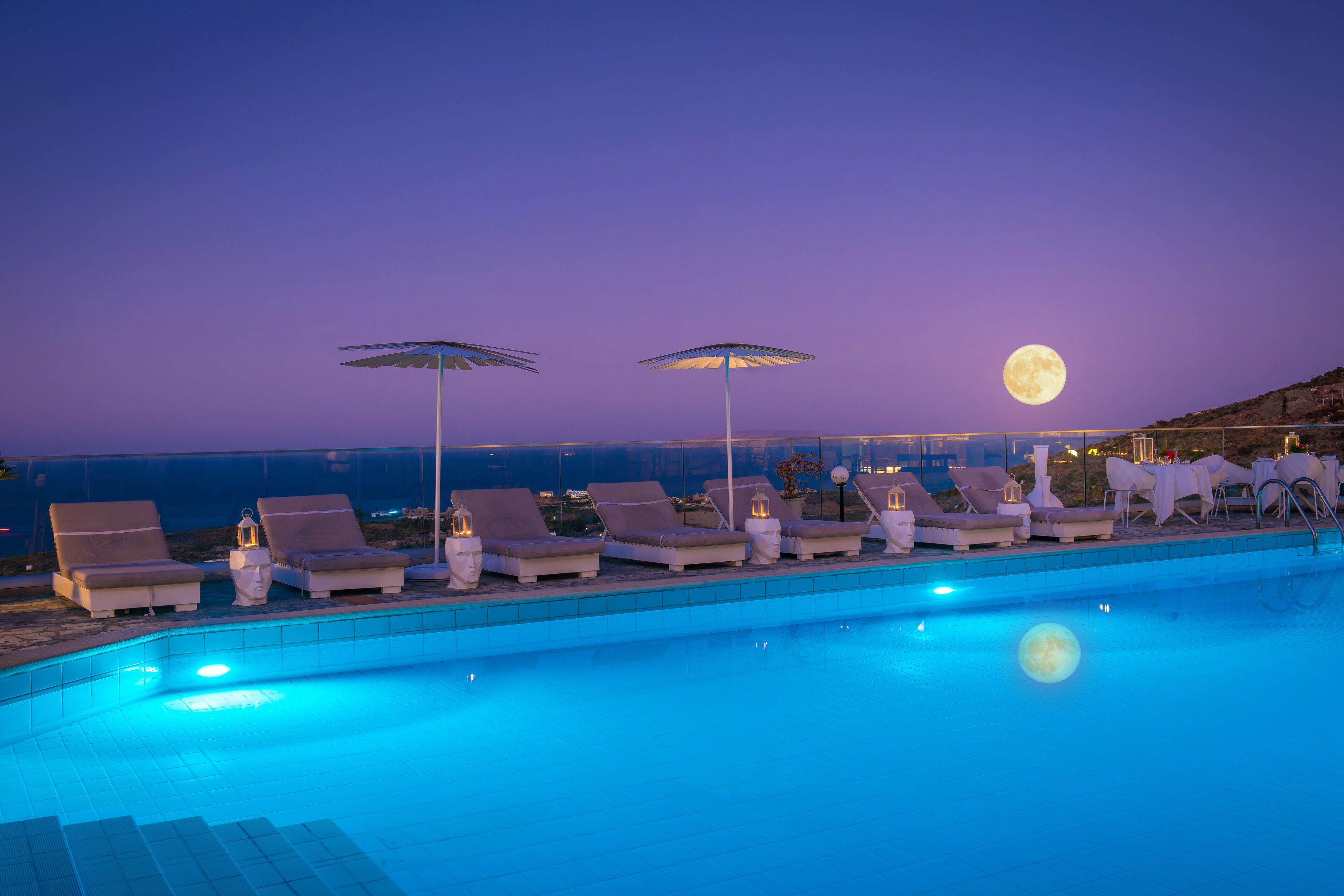 Ein Poolbereich mit Liegestühlen und Sonnenschirmen, ein Mond am Himmel und Blick auf das Meer.