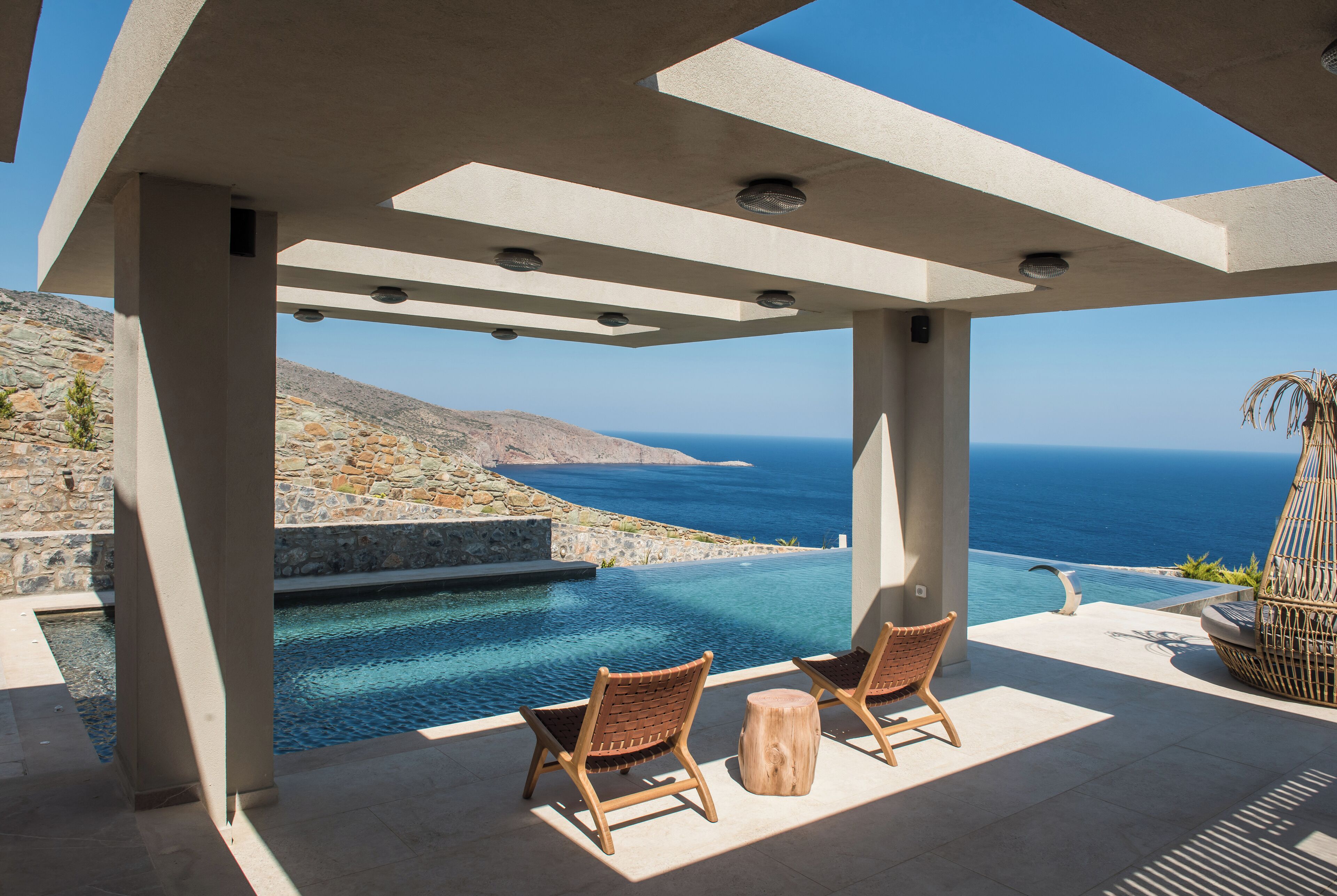 Un espace extérieur moderne, aménagé avec deux fauteuils en osier et un tabouret en bois, offrant une vue sur une piscine et un paysage magnifique de l’océan.