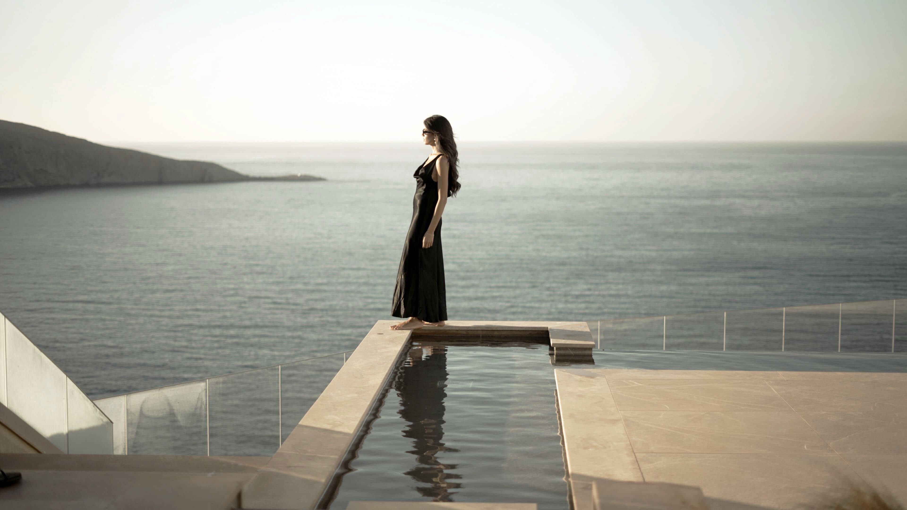 Une femme se tient au bord d’une piscine à débordement, d’où l’on a une vue imprenable sur la mer.