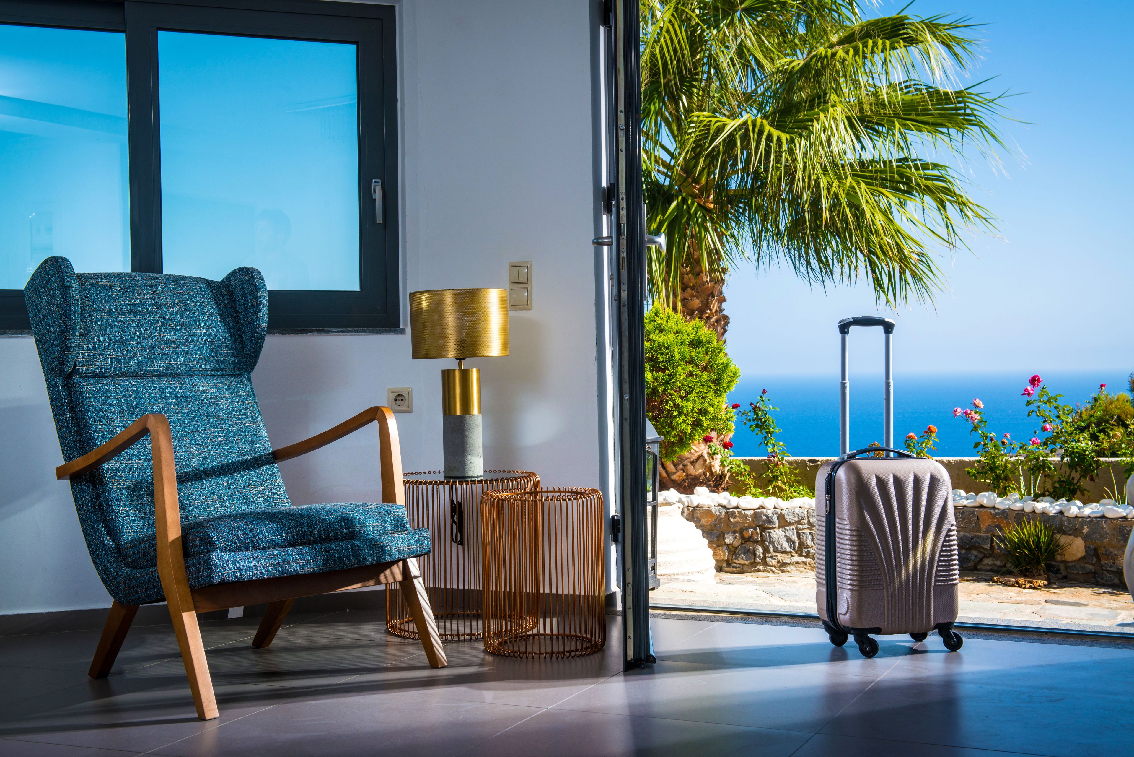 A room with a blue chair, a lamp, and a suitcase near a glass door with a view of the ocean.