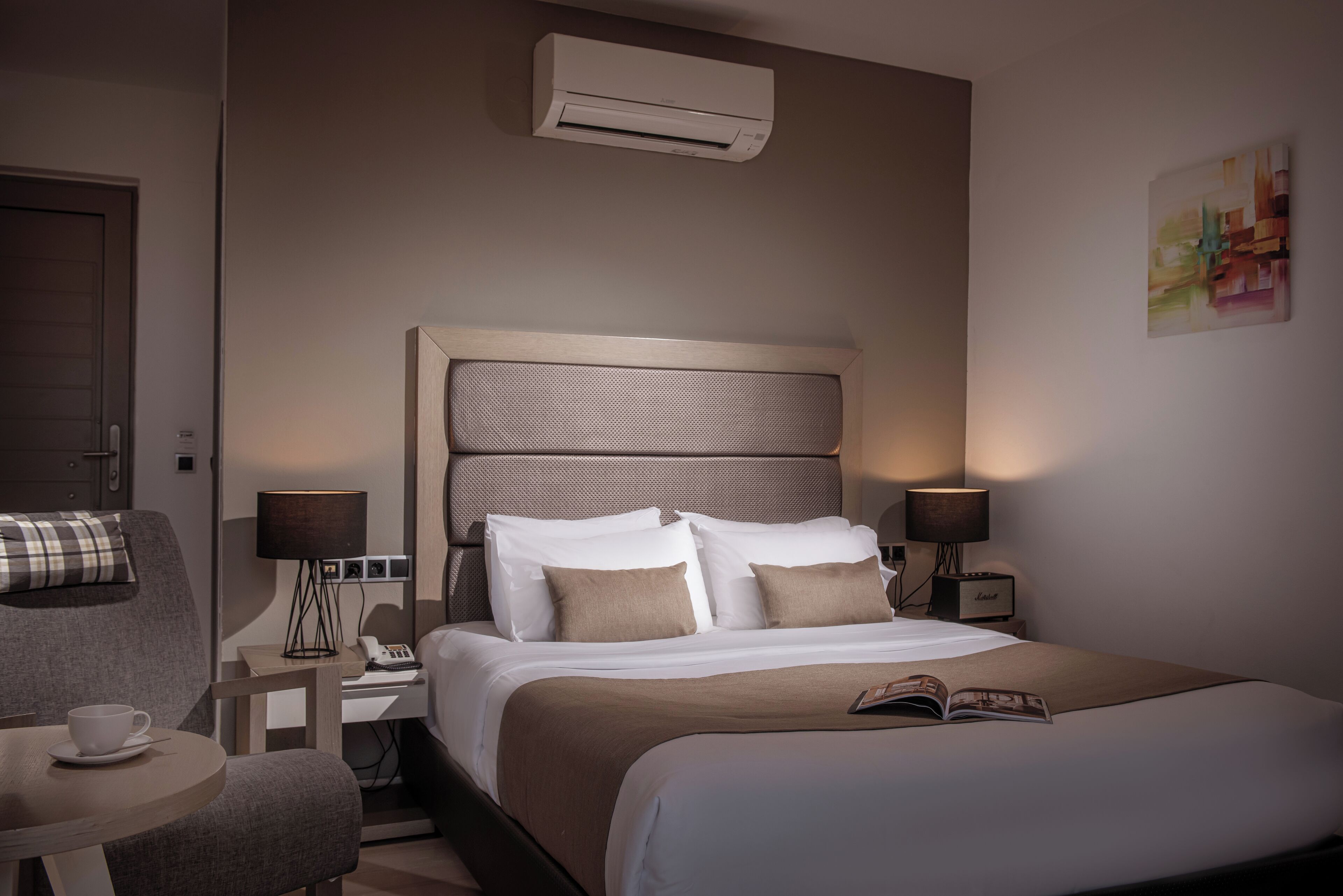 A hotel room with a large bed, a bedside table with a lamp, a chair, and an air conditioning unit on the wall.