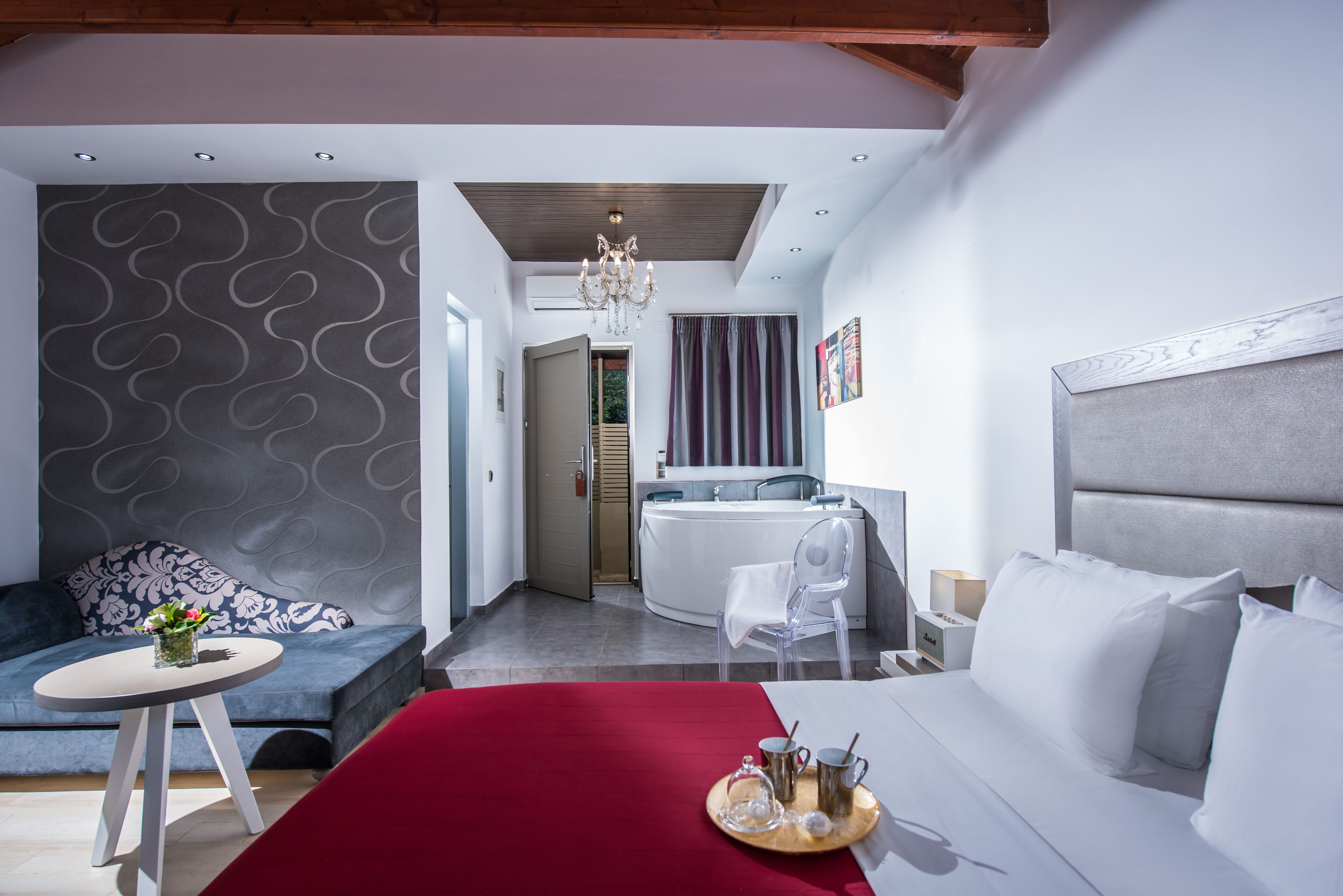 Una habitación de hotel moderna con una cama, una pequeña mesa redonda con una bandeja, un sofá, una silla y un baño visible a través de una puerta abierta.