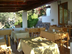 An outdoor dining area with tables and chairs, stone walls, and greenery in the background. - Albergo Villa Diana (Lipari)