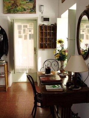 A dining area with a wooden table, chairs, a lamp, and a mirror. There is a door with a window, a wall-mounted key rack, and a painting above the door. - Albergo Villa Diana (Lipari)