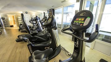 A gym with multiple cardio machines, including ellipticals and a stationary bike, with large windows providing natural light.