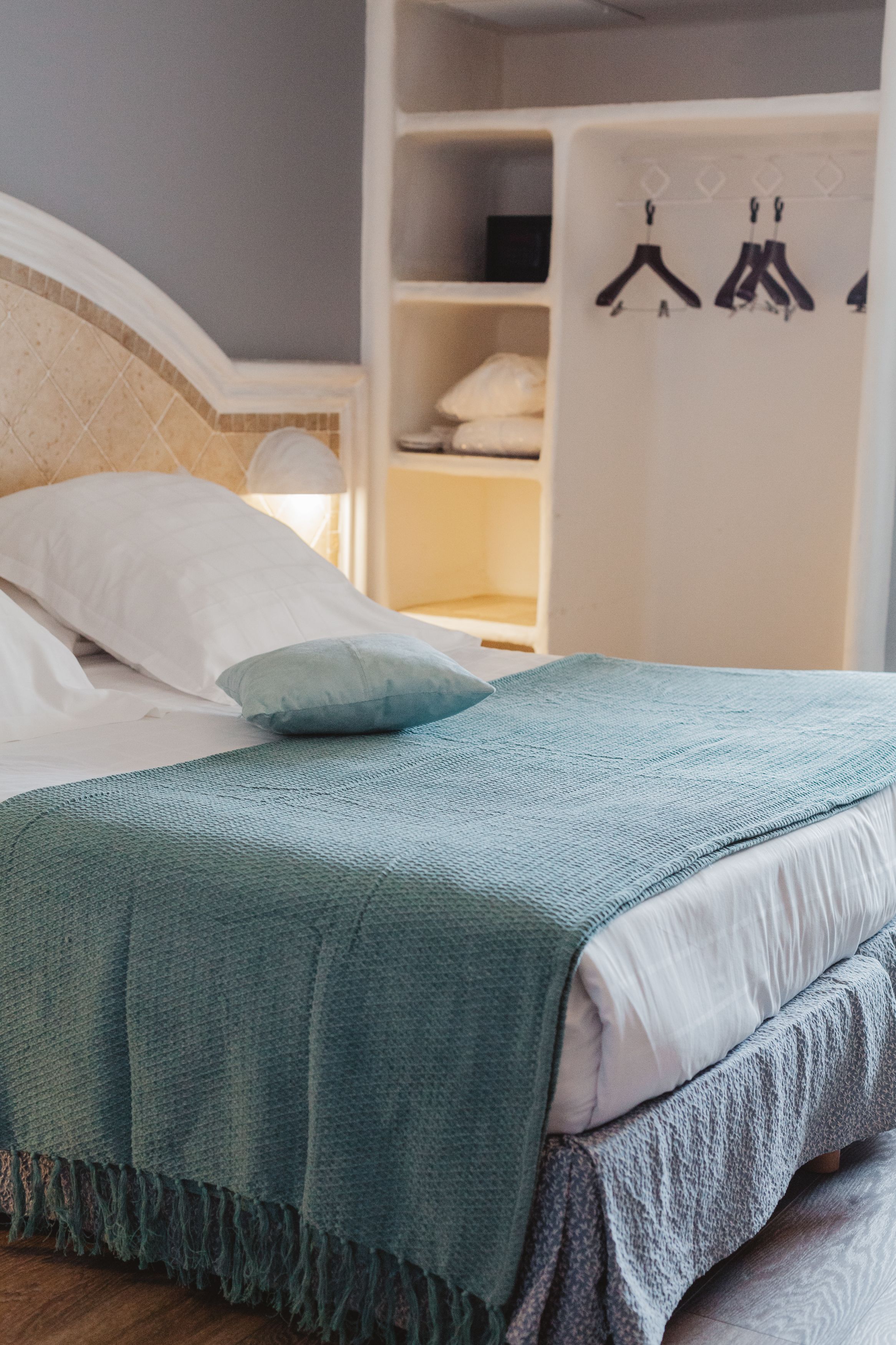 Een net opgemaakt bed met een blauwe deken en kussens, een witte kledingkast met hangers en een houten nachtkastje.