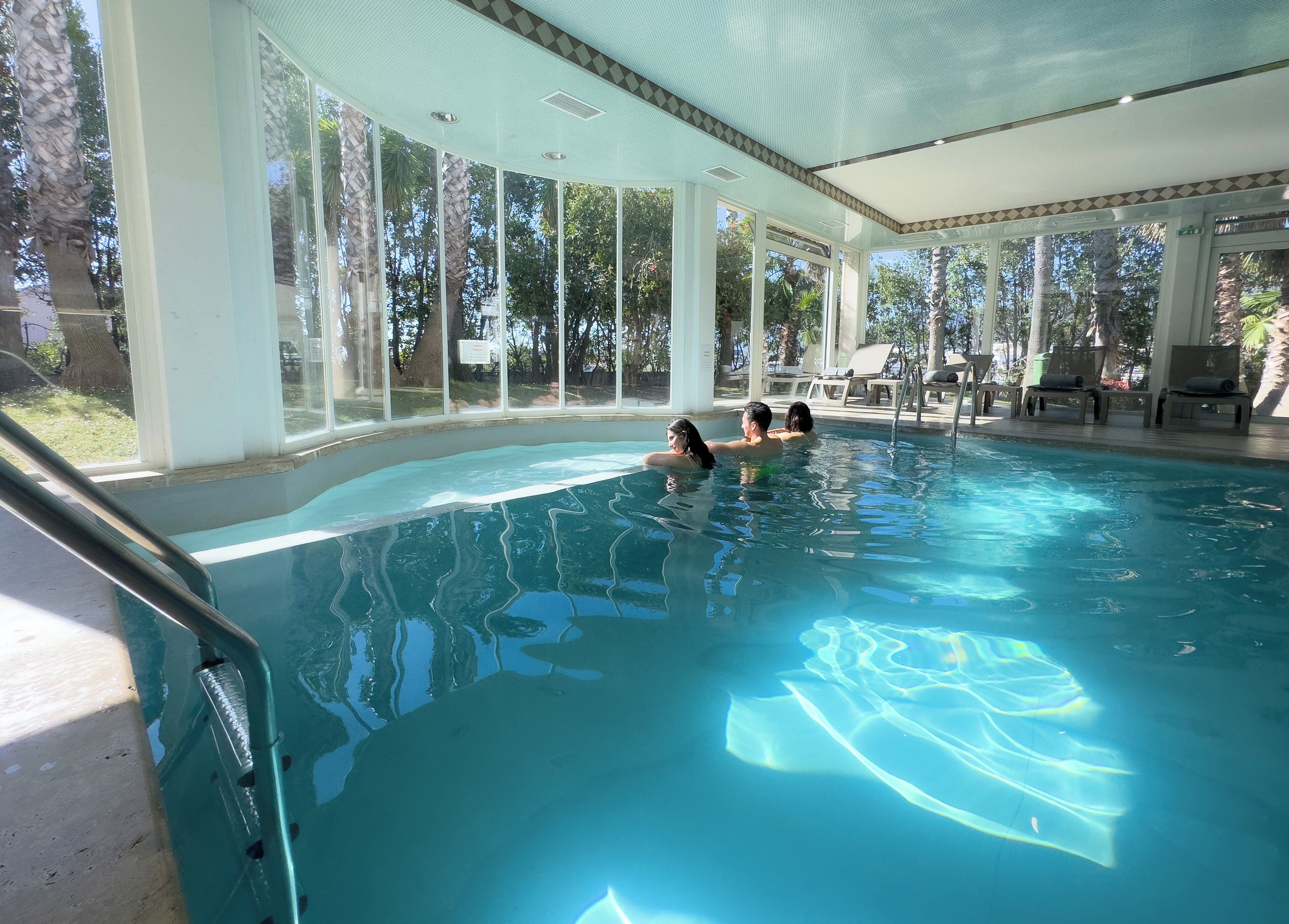 Una piscina interior con paredes de vidrio transparente, rodeada de sillas de descanso y vistas a árboles.
