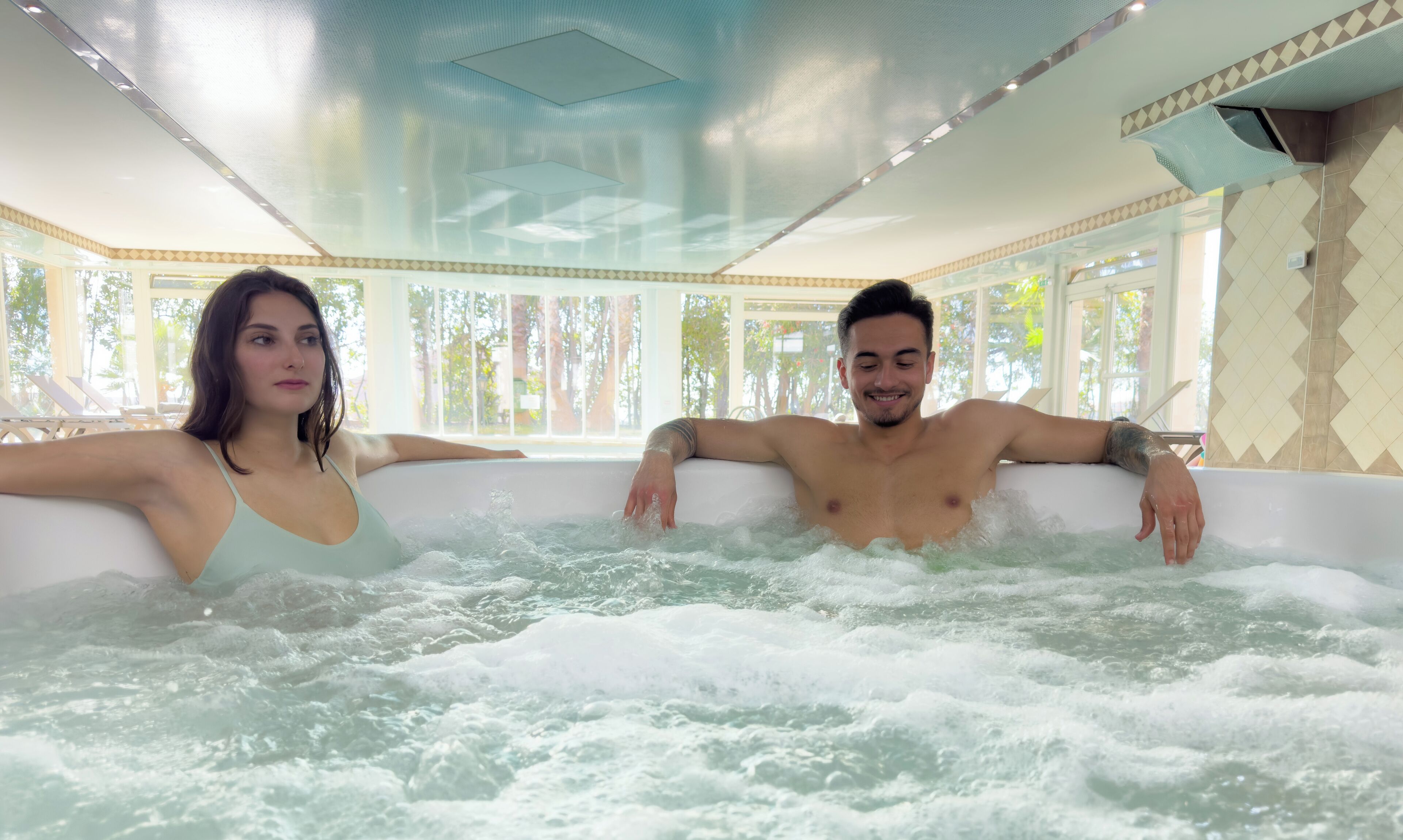 Two people relaxing in a hot tub with a view of trees through large windows.
