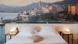 A bed with white linens and a view of a coastal town with buildings and a lighthouse. - Hôtel Spa Restaurant l'Ostella (Bastia)