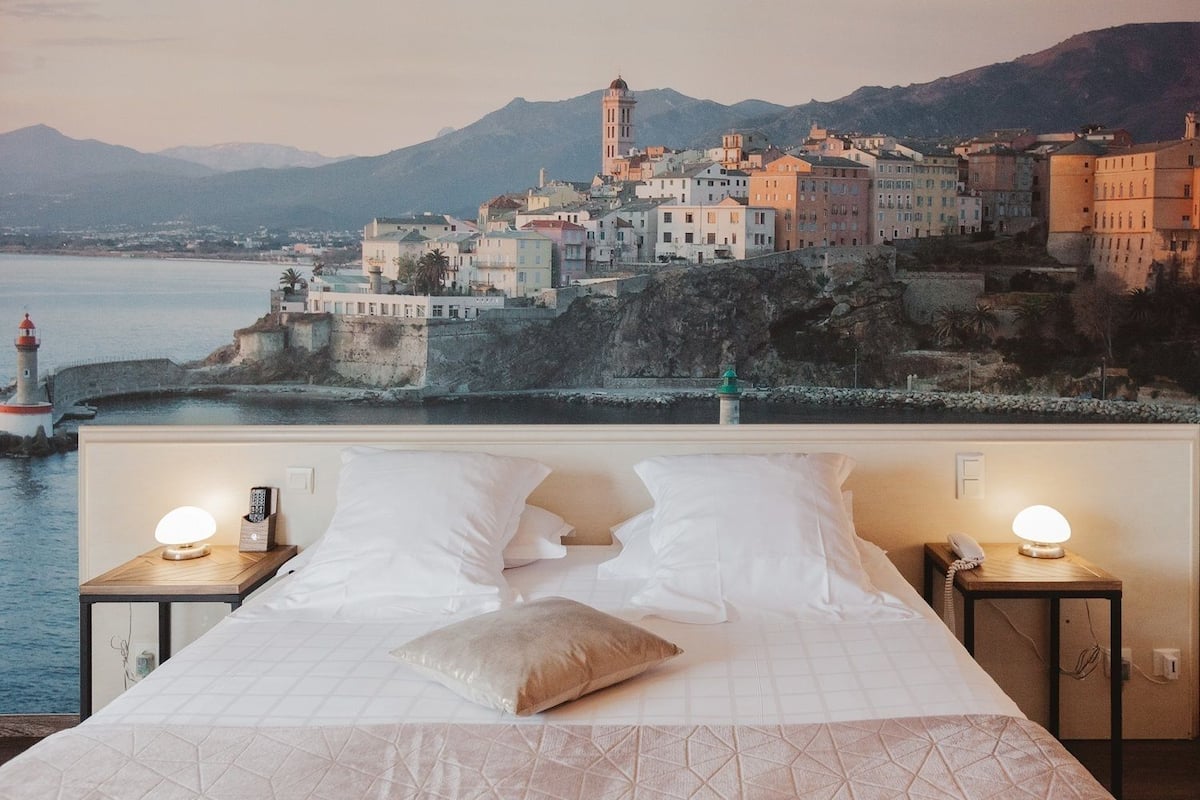 A bed with white linens and a view of a coastal town with buildings and a lighthouse.