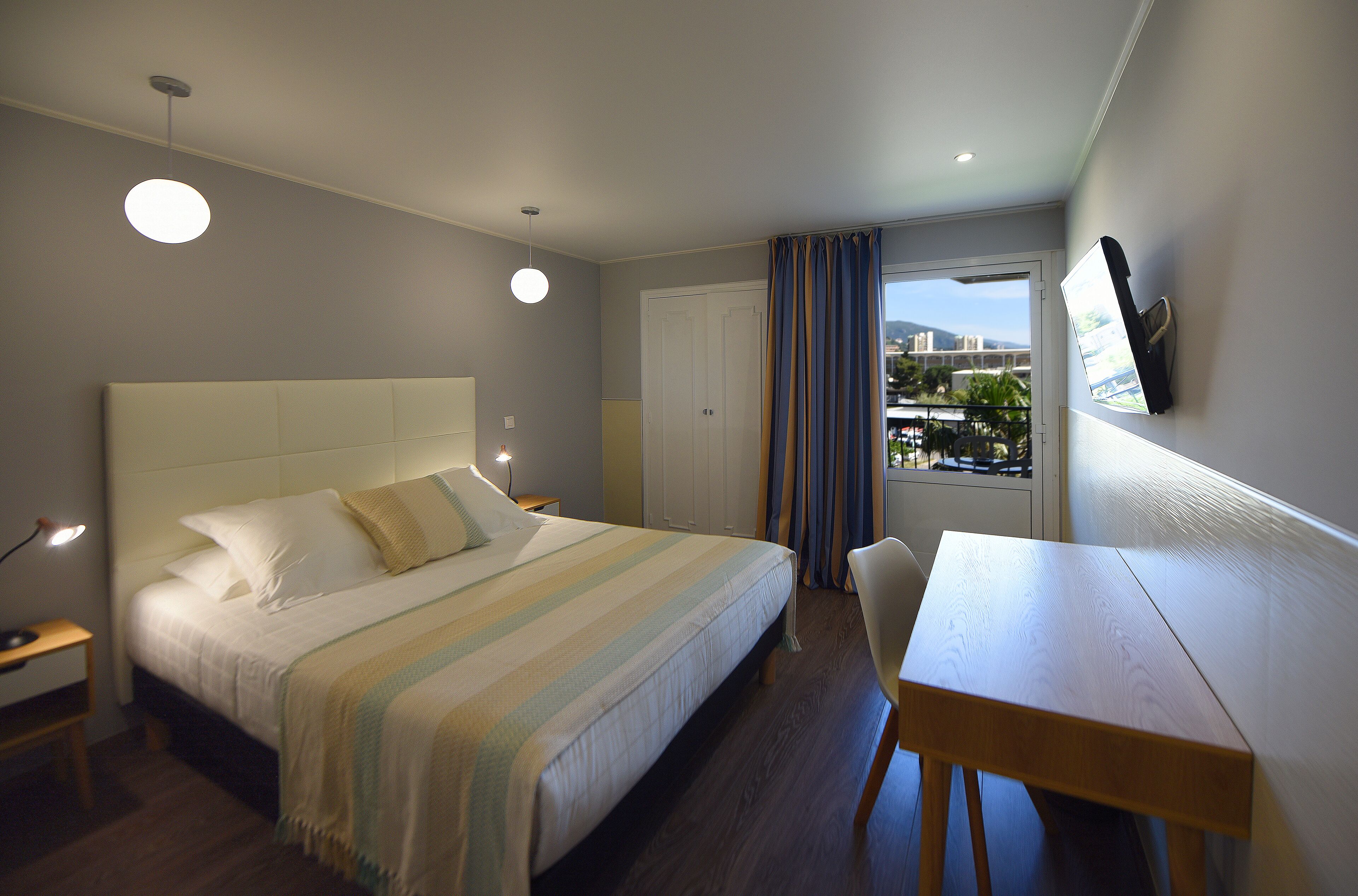 A hotel room with a bed, bedside table, desk, chair, and a TV mounted on the wall.