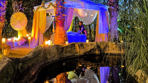 An outdoor event setup with a canopy, tables, and chairs, surrounded by palm trees and a stone bridge. - Hôtel Spa Restaurant l'Ostella (Bastia)