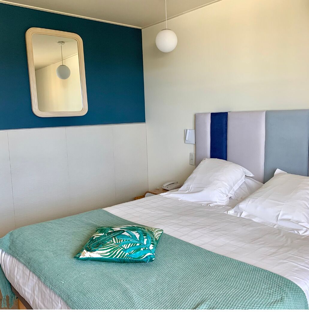 A hotel room with a bed, a bedside table, a mirror, and a wall-mounted light fixture.
