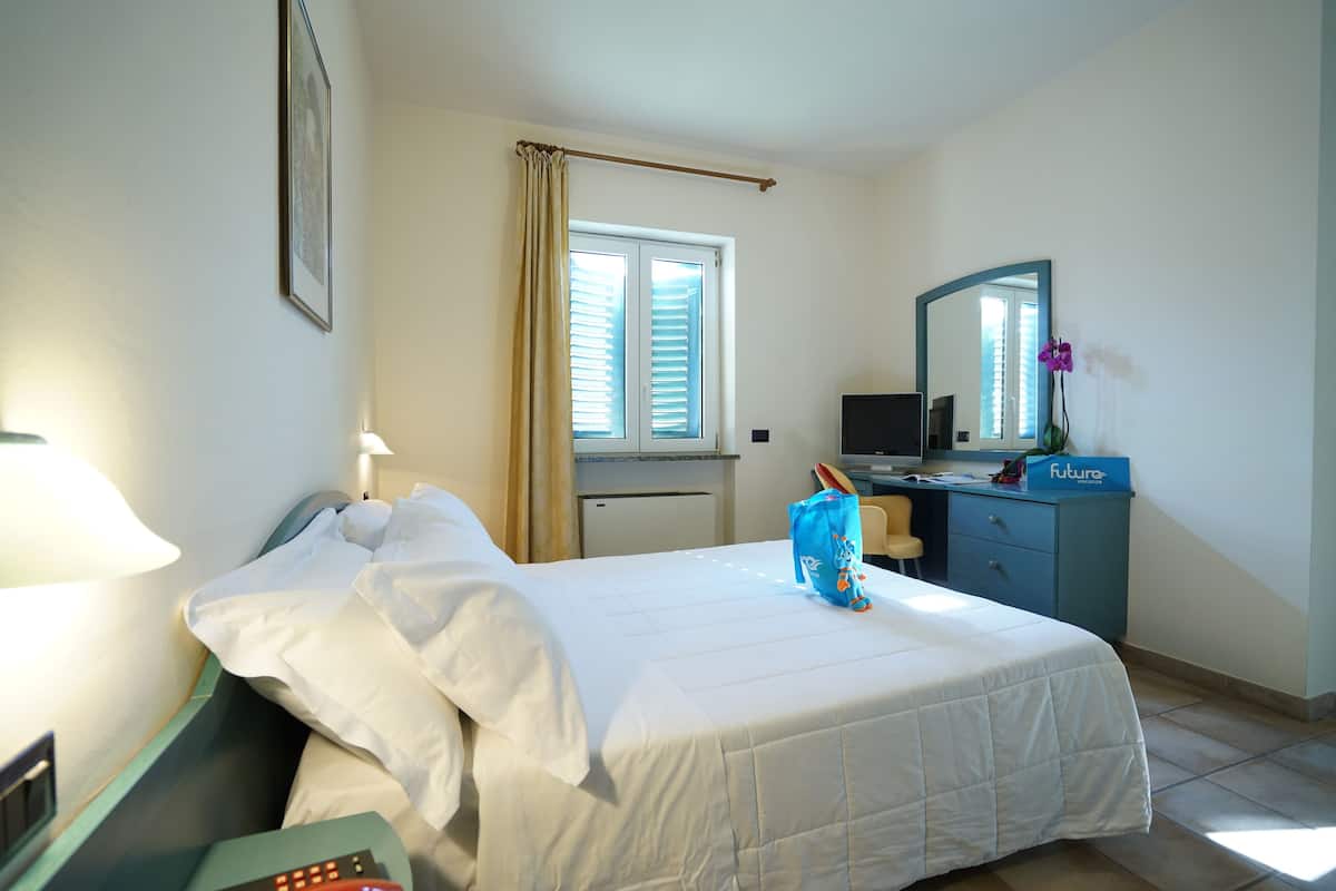 A hotel room with a bed, a desk, a chair, a desk lamp, a mirror, a window with curtains, and a small refrigerator.