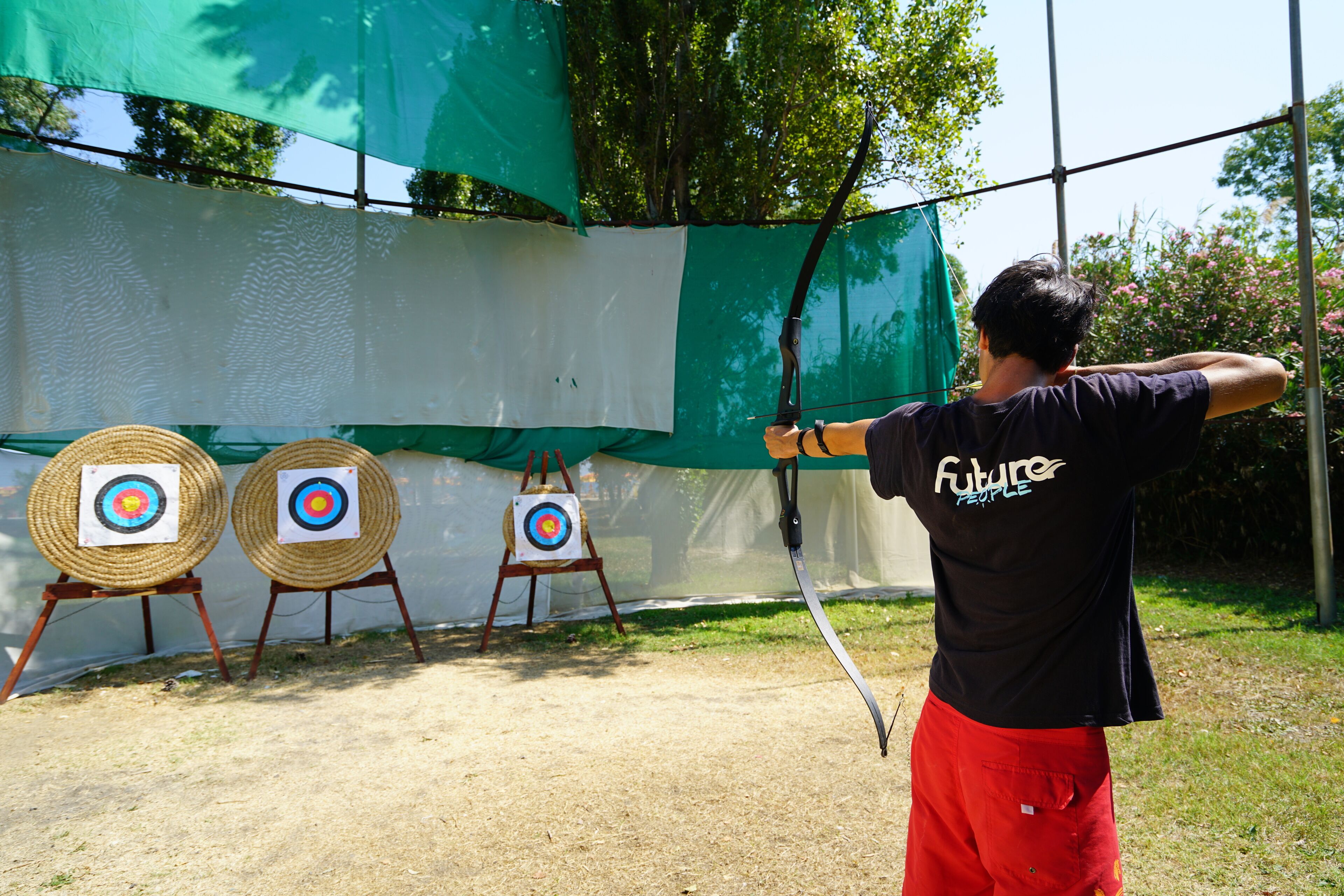 An indoor archery range with targets and a person in a black shirt and red shorts aiming a bow.