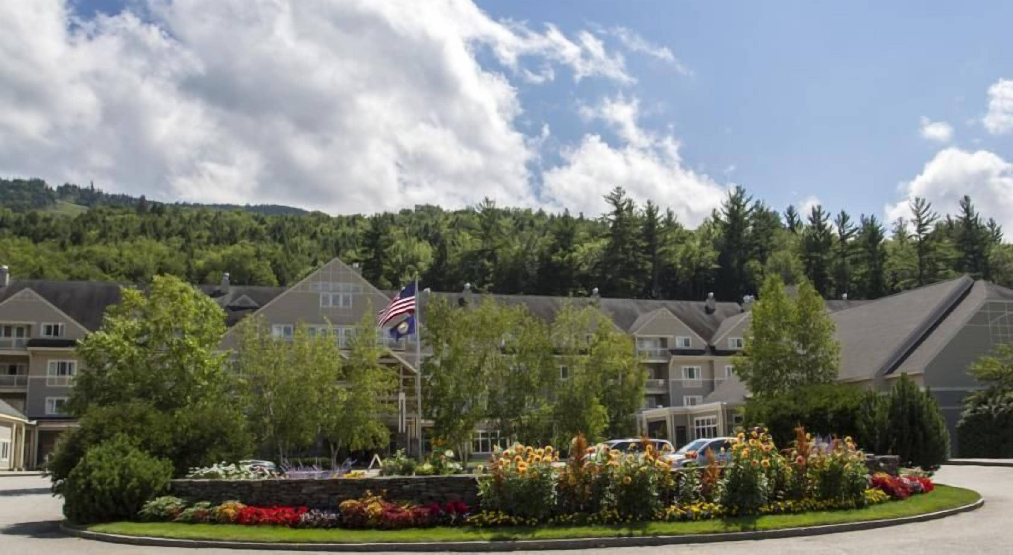 A hotel with a flagpole, surrounded by landscaped gardens and a forested hill in the background.