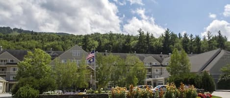 Un hotel con asta de bandera, rodeado de jardines ajardinados y una colina boscosa al fondo.