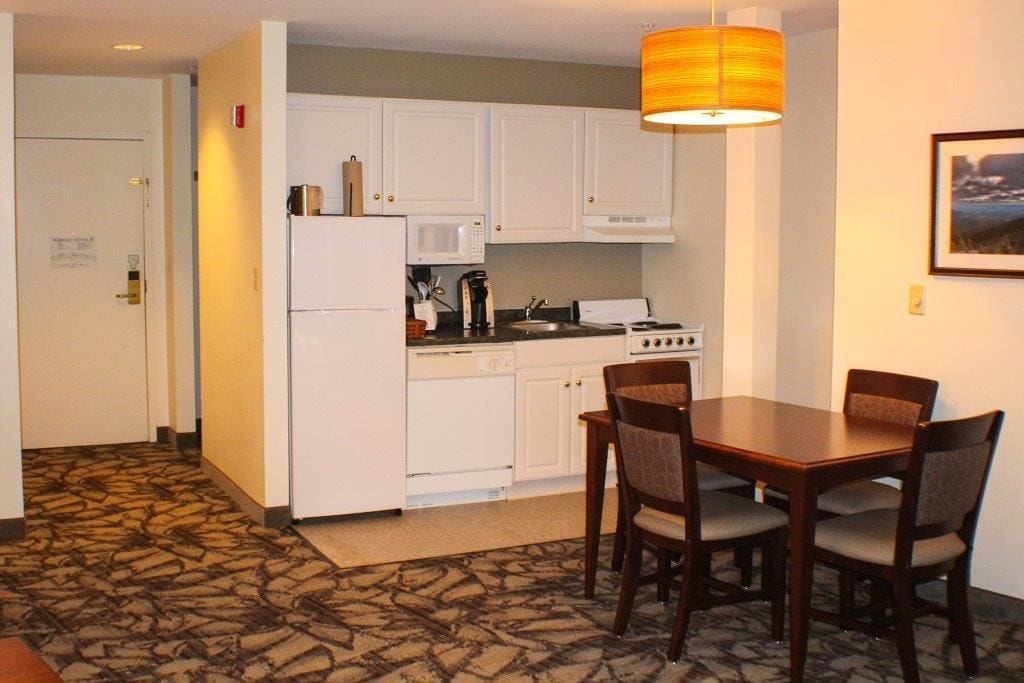 A compact kitchen with white cabinets, a refrigerator, a microwave, a coffee maker, and a stove. There is a dining table with four chairs.