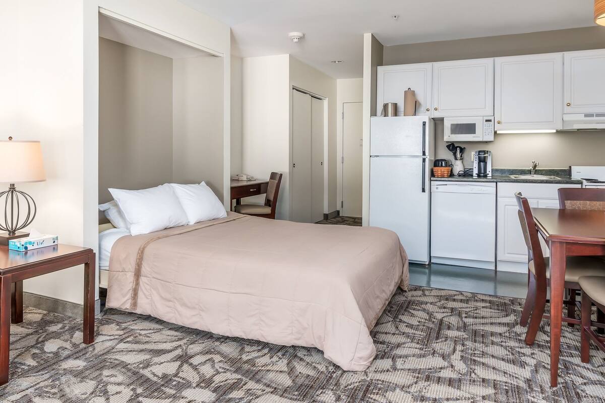 A hotel room with a bed, bedside table, lamp, and a kitchenette area with a microwave, refrigerator, and sink.