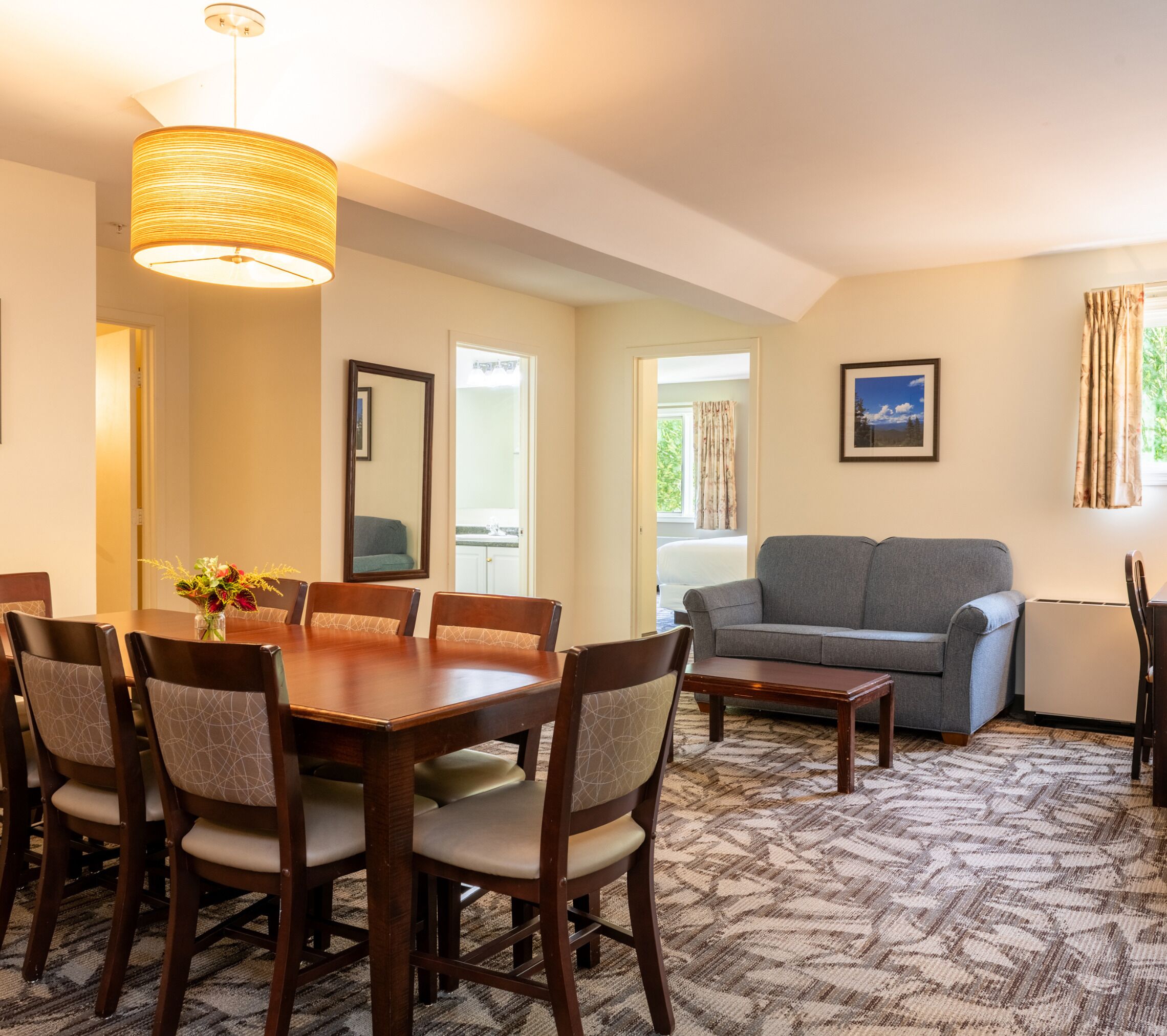 A hotel room with a dining area, a gray sofa, a large mirror, and a window with curtains.