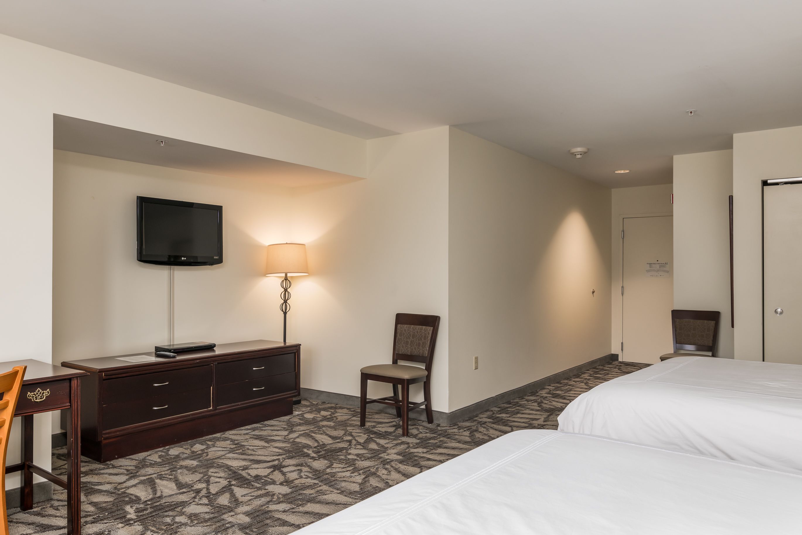 A hotel room with a bed, a desk with a lamp, a chair, and a TV mounted on the wall.