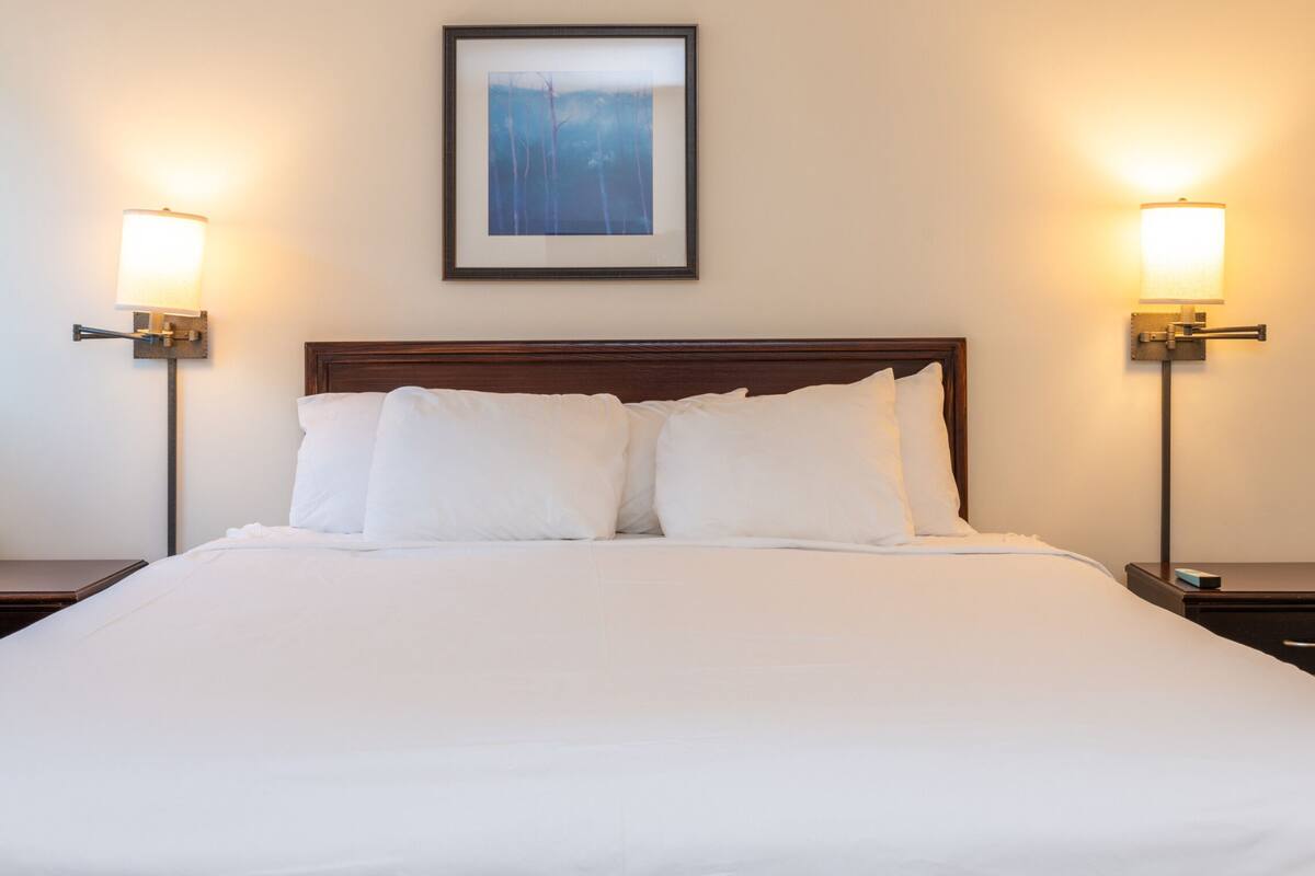 A neatly made bed with white linens, flanked by two bedside tables with lamps, and a framed picture above the headboard.