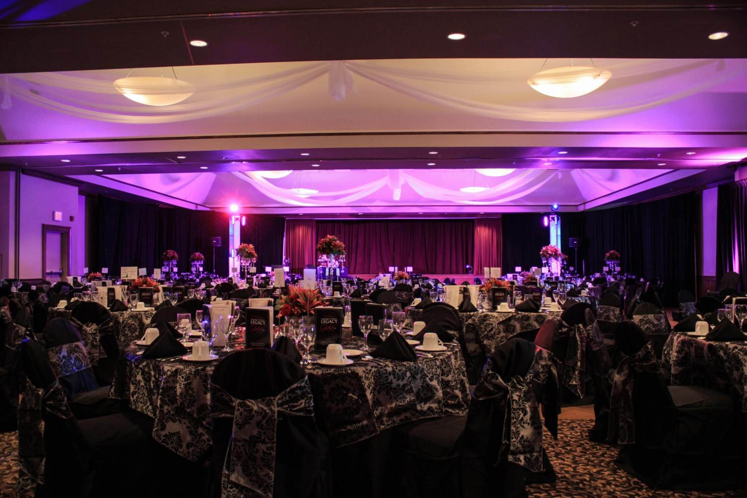 Een balzaal met ronde tafels gedekt voor een formeel evenement, met een podium en bloemdecoraties.