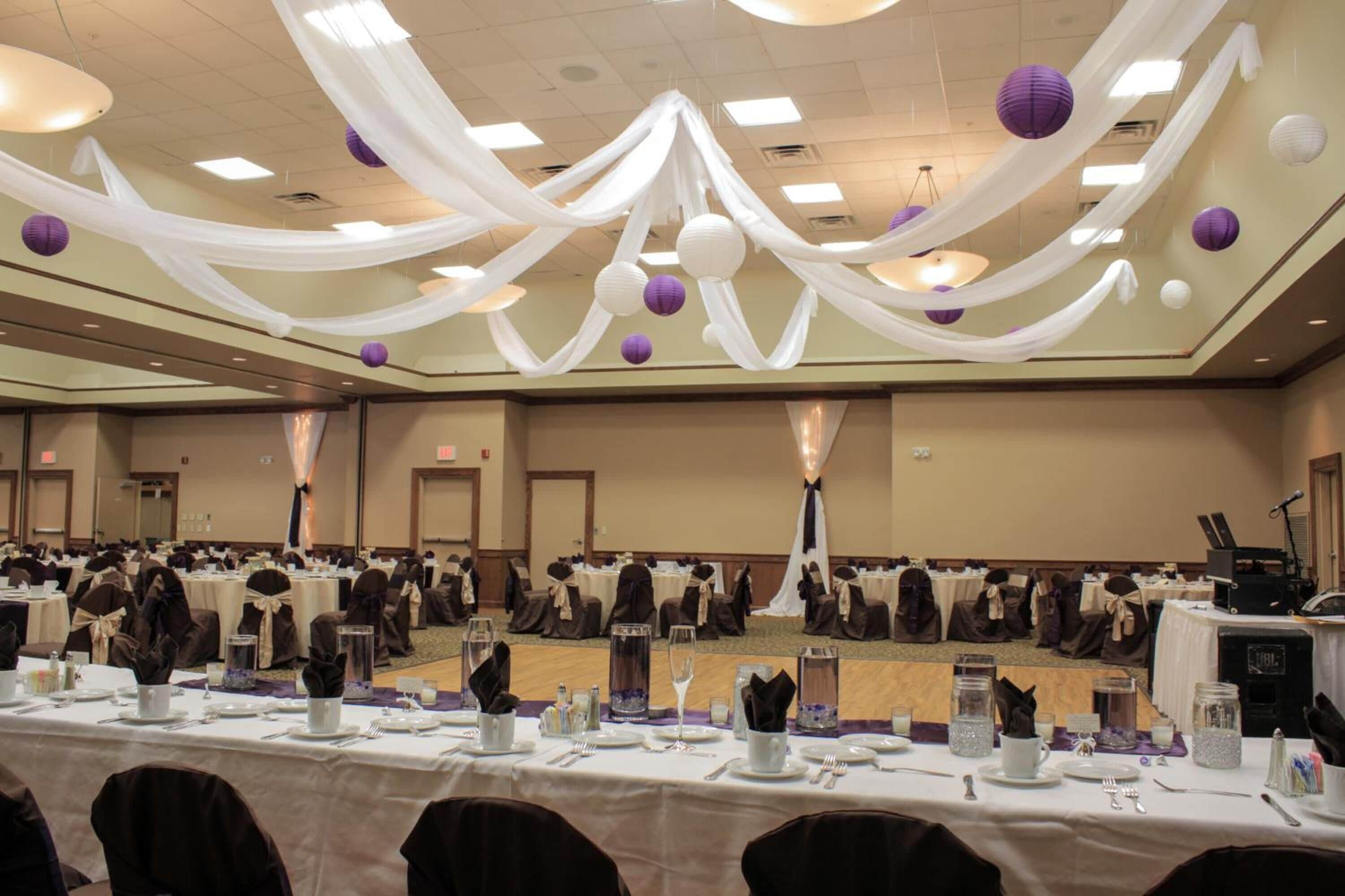 A banquet hall with tables set for a formal event, draped with white fabric and purple decorations.