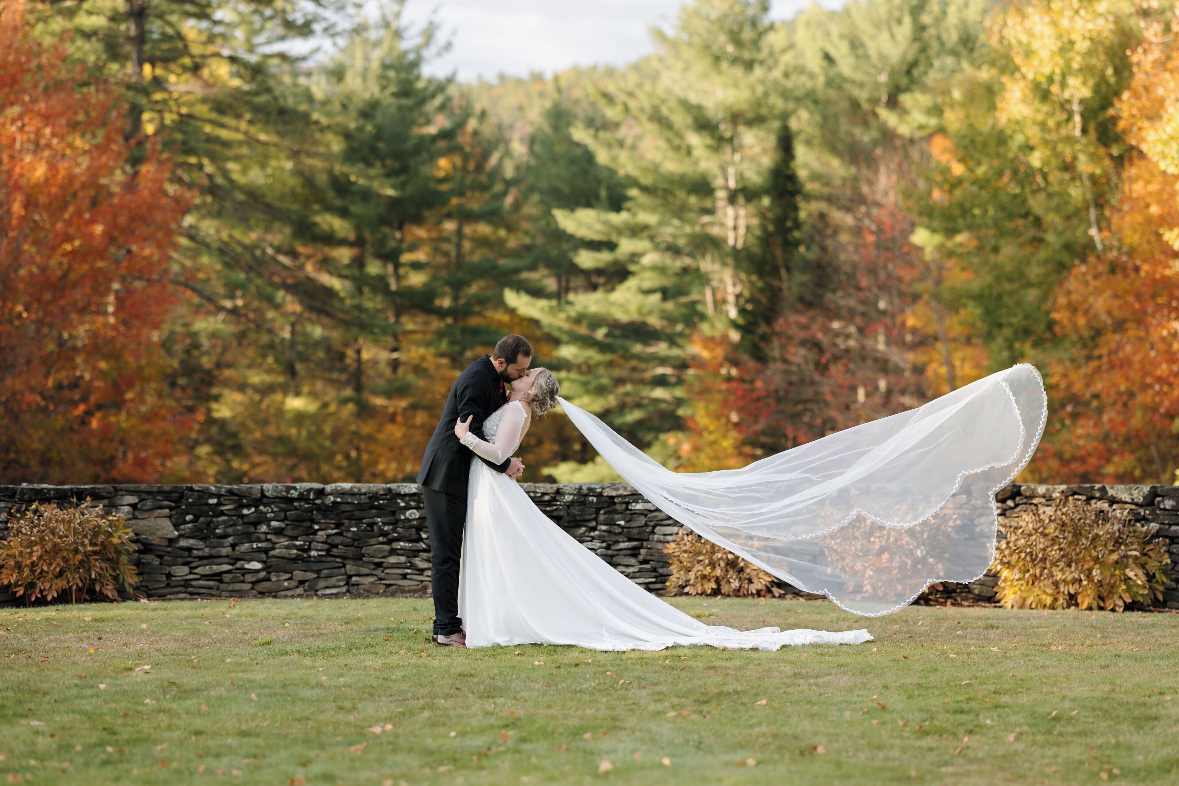 A couple embracing in a grassy outdoor area with a stone wall and autumn foliage in the background.