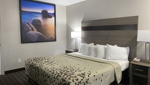A hotel room with a bed, a nightstand, a lamp, and a painting of a coastal scene. - High Country Lodge (South Lake Tahoe)