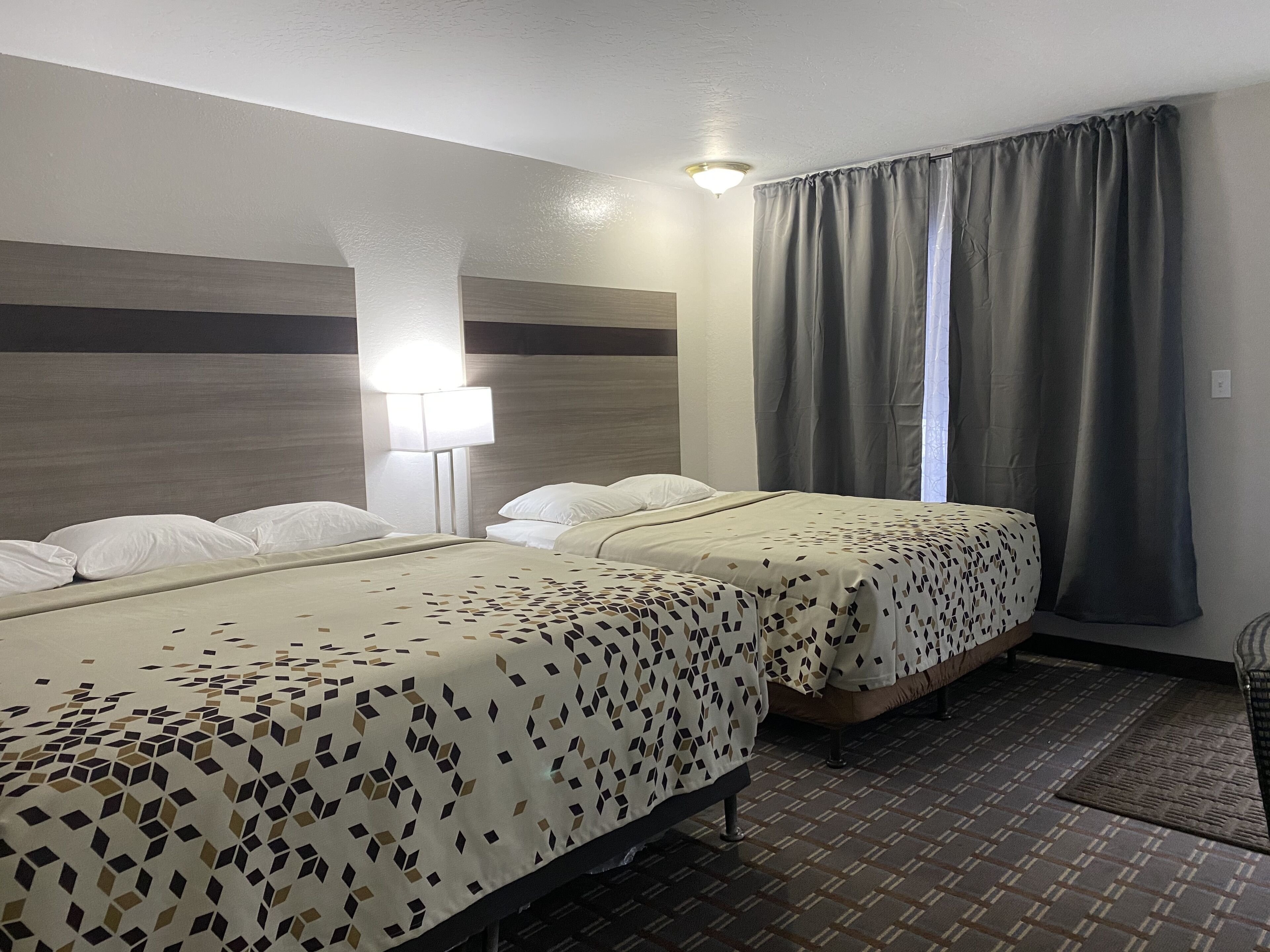 A hotel room with two beds, a nightstand, a lamp, and a window with curtains.