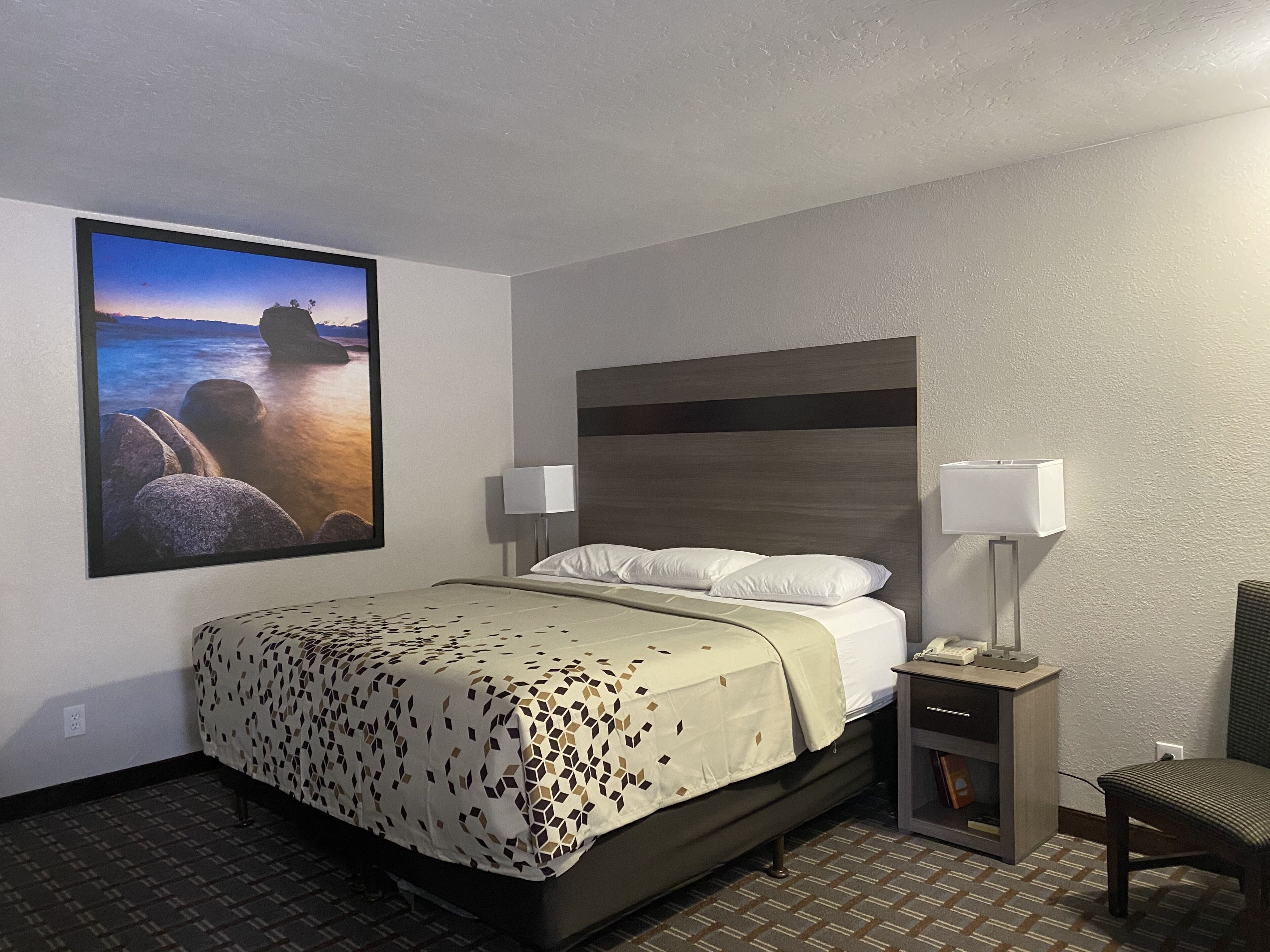 Ein Hotelzimmer mit einem Bett, zwei Nachttischlampen, einem Sessel, einem Nachttisch und einem großen gerahmten Bild mit Strandmotiv.