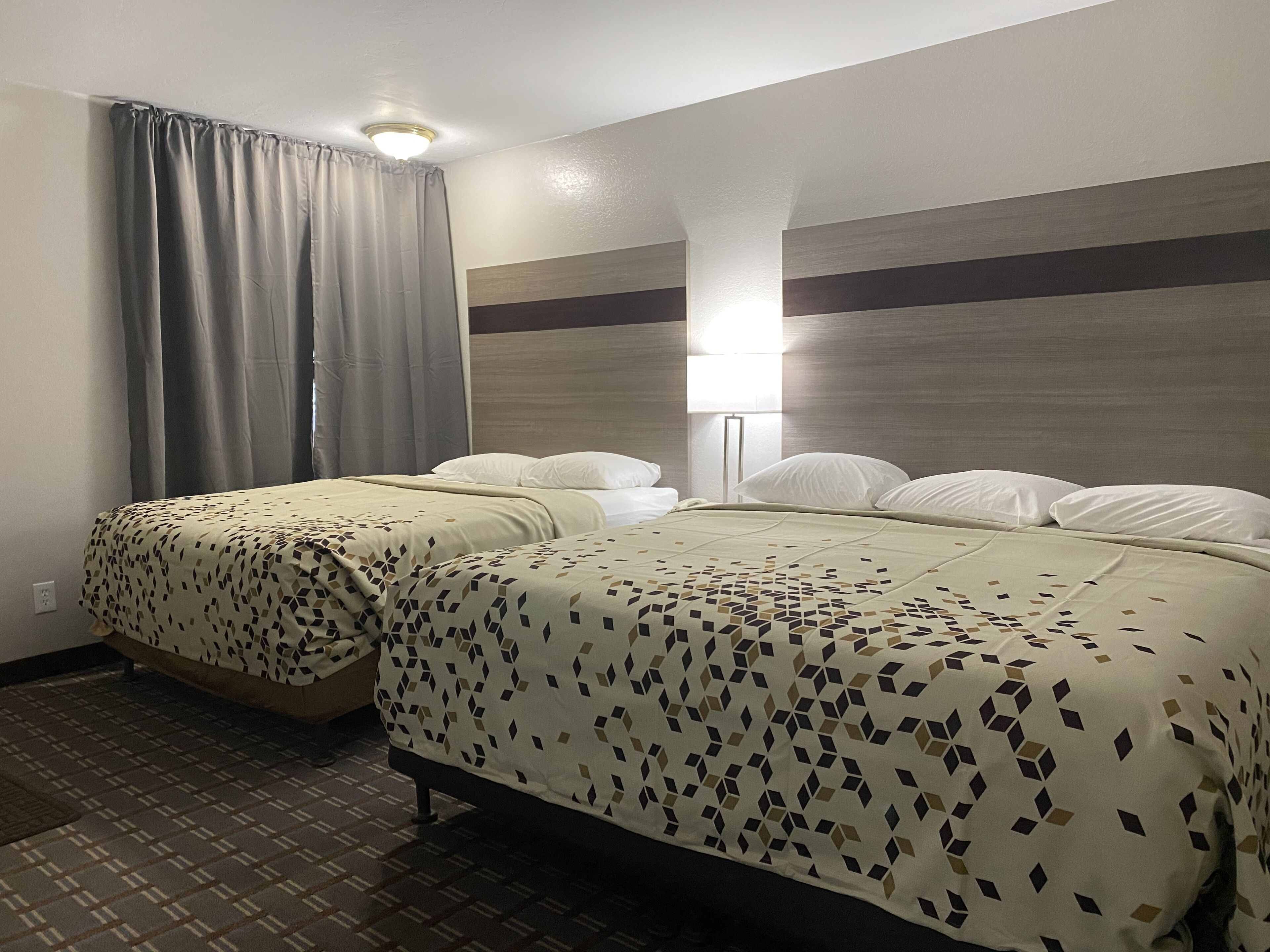 Hotelkamer met twee bedden, een hoofdbord met lamp en een nachtkastje, en een raam met gordijnen.