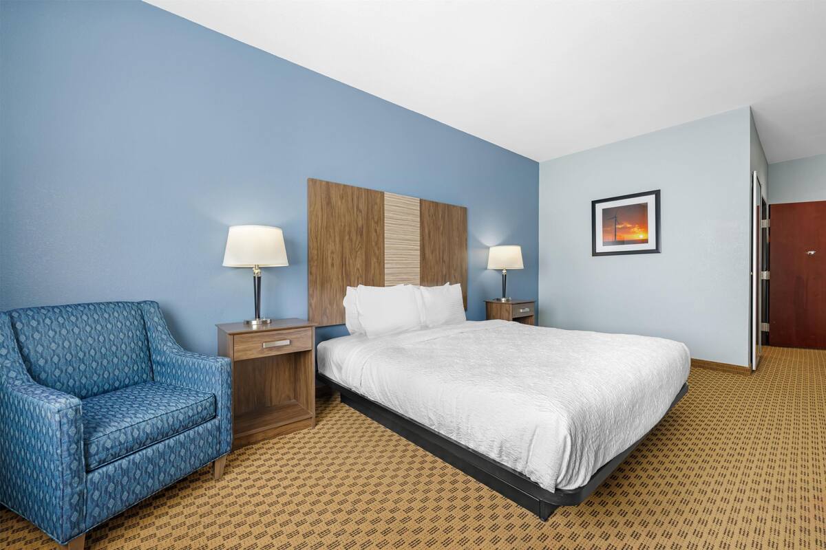 A hotel room with a bed, a blue armchair, two bedside tables with lamps, a wooden headboard, and a framed picture on the wall.