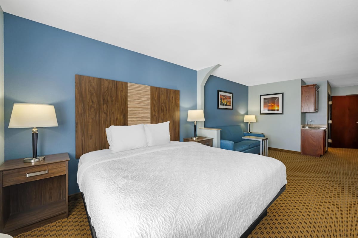 A hotel room with a large bed, two bedside tables with lamps, a desk with a chair, a small bathroom, and a wooden headboard.