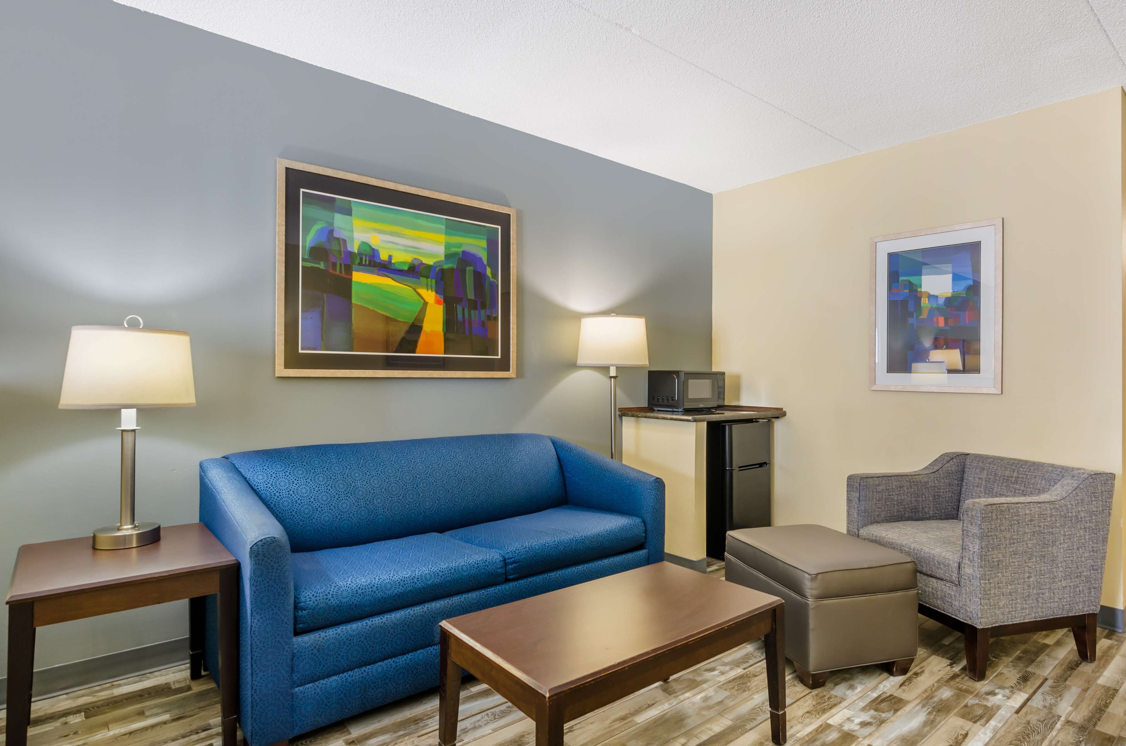 A hotel room with a blue sofa, a grey armchair, a wooden coffee table, two lamps, a microwave, and a small cabinet.