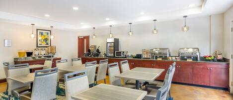 Un comedor de hotel con mesas y sillas, una zona de cocina con electrodomésticos e iluminación colgante.