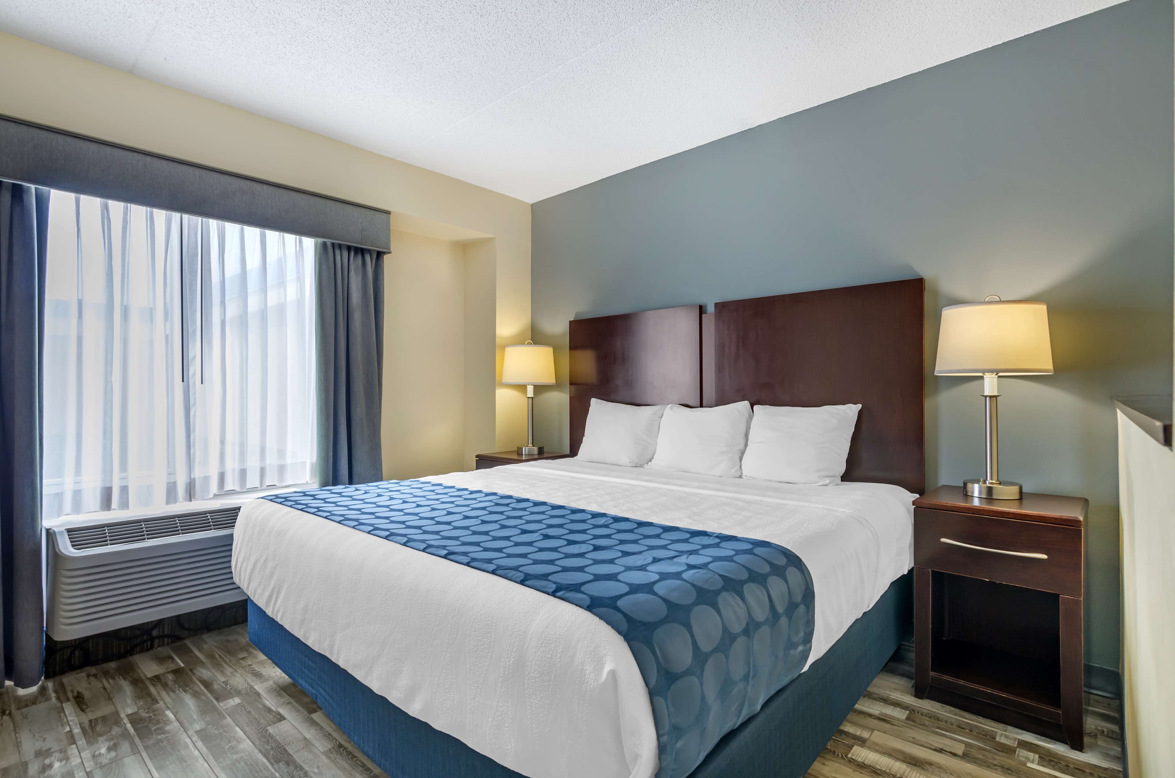 A hotel room with a large bed, two bedside tables with lamps, a window with curtains, and a wall-mounted air conditioner.