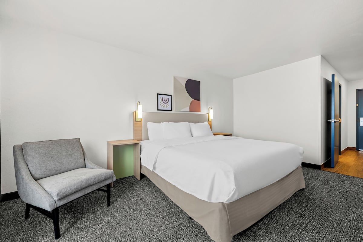 A hotel room with a bed, a chair, a nightstand, and a wall-mounted lamp.