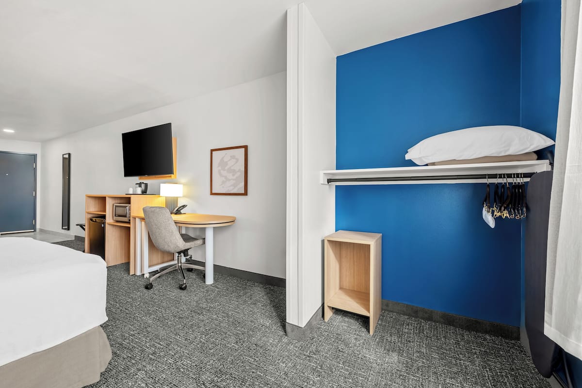 A hotel room with a bed, desk, chair, TV, and a blue accent wall.
