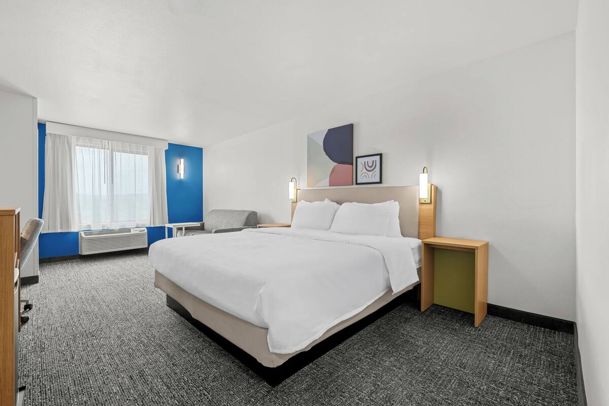 A hotel room with a large bed, a desk, a chair, a window with curtains, and a wall-mounted artwork.